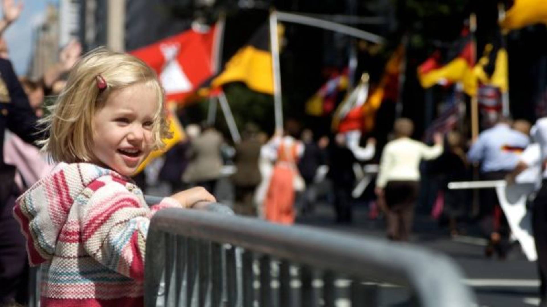 little girl in hoodie holding onto metal barrier and looking at street parade